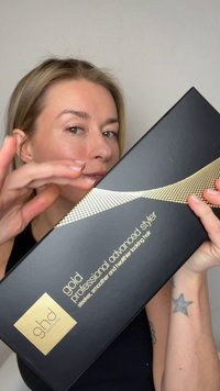 Kvinde med blondt hår, der holder en sort og guldbox mærket "gold professional advanced styler" til glattere, mere glansfuldt og sundere udseende hår.