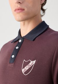 Tricou polo mahon cu guler bleu închis. Prezintă butoane albe și un logo brodat „Les Deux” sub formă de scut pe piept. Textură netedă.