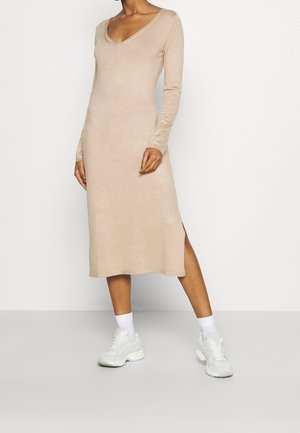 Frau trägt ein beiges Midi-Kleid mit langen Ärmeln und Seitenschlitz, weiße Socken und weiße klobige Sneaker, steht vor einem einfarbigen hellen Hintergrund.