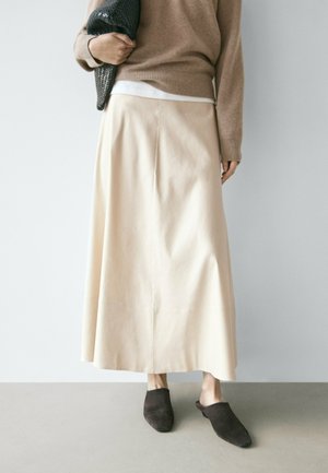 Persoon met een beige lange rok, een bruine trui, zwarte slip-on schoenen en een gestructureerde zwarte clutch vastgehouden tegen een effen achtergrond.