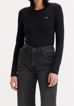 Femme portant un haut noir à manches longues Levi's rentré dans un jean noir délavé taille haute, debout devant un fond uni.