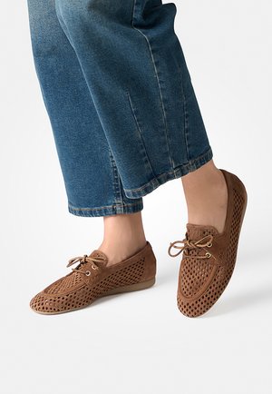 Brown perforated lace-up loafers worn with cropped blue denim jeans, shown from mid-calf down on a white background.