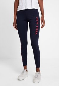 Leggings navy en matériau extensible avec "NEW YORK" imprimé en rouge verticalement sur une jambe ; assortis à des baskets blanches.
