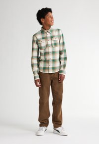 Kostkovaná košile v zelených, modrých a hnědých tónech s límečkem na knoflíky a dvěma předními kapsami, spárovaná s hnědými kordovým kalhotami a bílými teniskami.