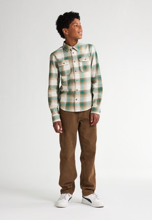 Geruite shirt in groen, blauw en bruin tinten met een knoopsluiting bij de kraag en twee voorzakken, gecombineerd met een bruine corduroy broek en witte sneakers.