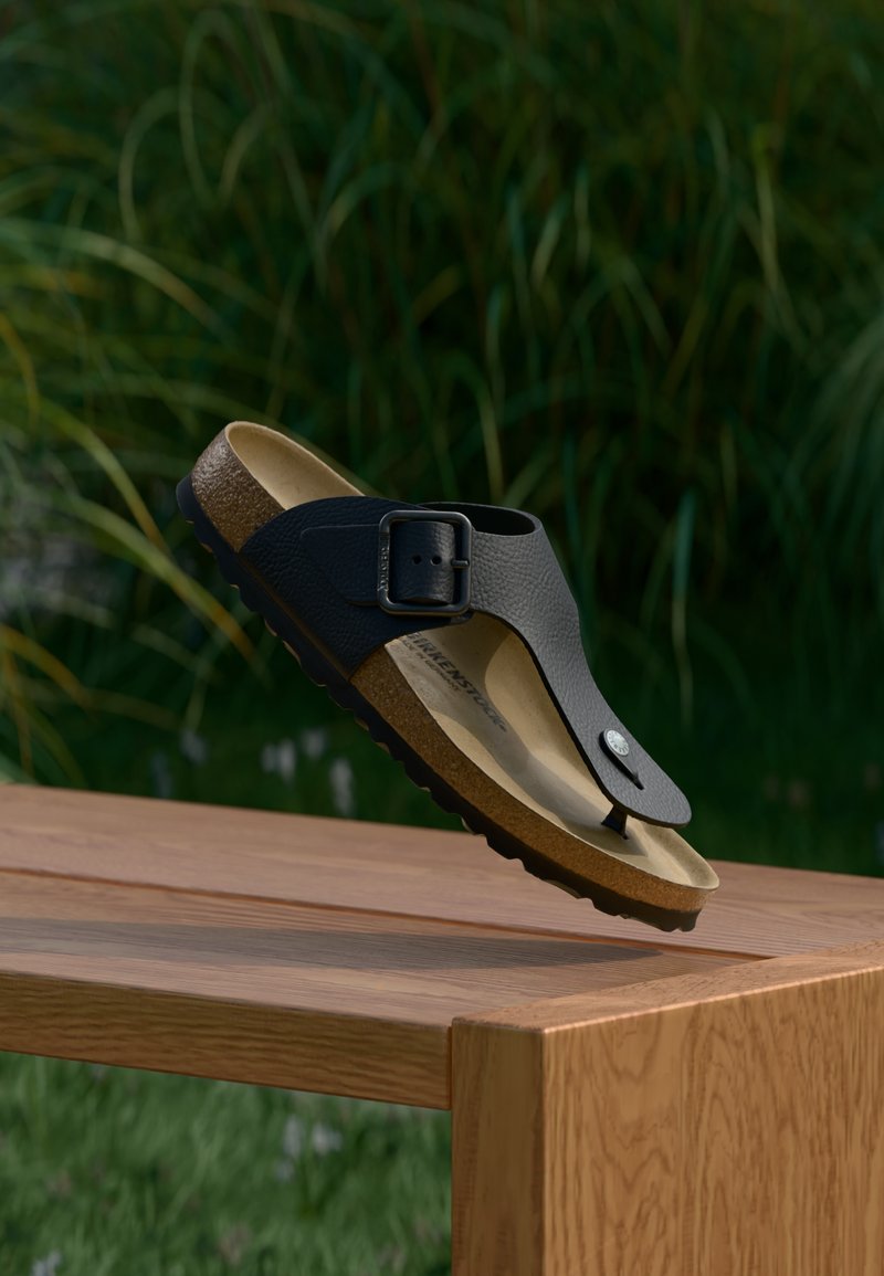 Μαύρο σανδάλι Birkenstock με αγκράφα και φελλό σόλας, αιωρούμενο πάνω από ένα ξύλινο παγκάκι σε εξωτερικό χώρο με φόντο πράσινη βλάστηση.