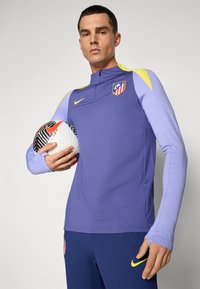 Lilla atletiktrøje med lange ærmer, gule accenter på skuldrene, prydet med Atlético Madrids logo og en Nike swoosh. Spilleren holder en fodbold.