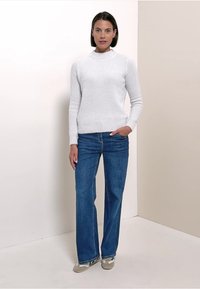 Maglione bianco in maglia con collo rotondo, indossato con jeans blu. Il maglione presenta un motivo a rilievo; la modella ha i capelli corti e si trova in posizione informale.