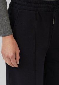 Lewa ręka spoczywa obok czarnych spodni dresowych z boczną kieszenią i sznurkiem w talii, na sobie ma szary, długi rękaw.