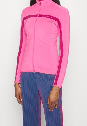 Kobieta w dopasowanej różowej kurtce na zamek z ciemnoróżowymi paskami oraz w niebieskich spodniach z czerwonymi pionowymi prążkami.