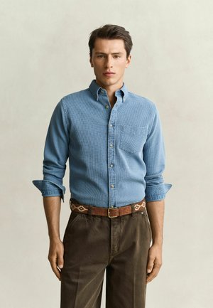 Homme portant une chemise bleu clair à boutons avec les manches retroussées, un pantalon marron et une ceinture marron à motifs, debout devant un fond uni.