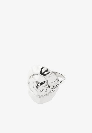 Anillo de metal plateado con un diseño grande, irregular, ondulado y parecido a una flor en la parte superior, que refleja la luz sobre un fondo blanco.