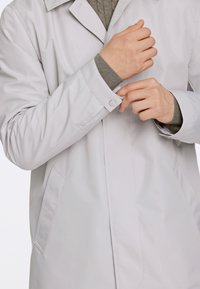 Cappotto impermeabile di colore grigio chiaro, con colletto classico, chiusure a scatto e tasche laterali. Polsini a costine visibili sotto le maniche del cappotto.
