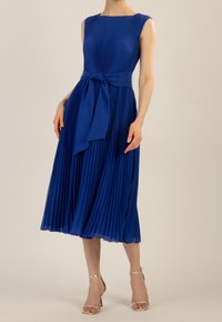 Blue pleated dress with a fitted bodice and a waistband tied into a bow. Smooth texture, knee-length, and complemented by silver heels.