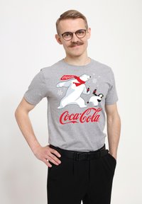 T-shirt grigia con grafica di un orso polare e di un pinguino, con branding rosso di Coca-Cola. Maniche corte e vestibilità standard. Pantaloni neri indossati.