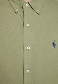 Světle olivově zelená košile na knoflíky s bílými knoflíky a malým vyšitým logem modrého polo hráče na hrudi.