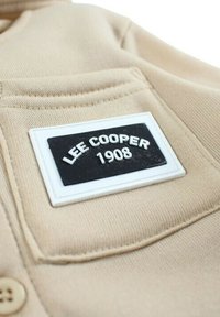 Tissu beige à surface texturée, détail de poche avec une étiquette noire portant l'inscription "LEE COOPER 1908" en lettres blanches.