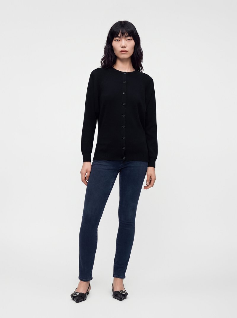 Femme portant un cardigan noir boutonné, un jean skinny bleu foncé et des ballerines noires pointues, debout sur un fond blanc uni.