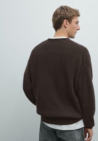 Bruine gebreide trui met een ribbelstructuur, relaxte pasvorm en ronde halslijn, gelaagd over een wit shirt, gestyled met grijze broek.