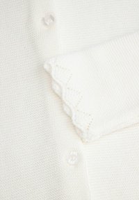 Biały sweter robiony na drutach z fakturowanej tkaniny, z falbaniastym mankietem i przezroczonym guzikiem.