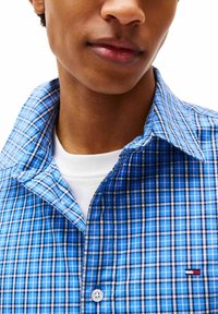 Primo piano di una persona che indossa una camicia a quadri blu sopra una maglietta bianca, mostrando parte del volto e del colletto.