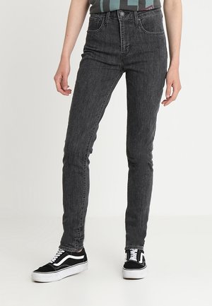 Schmal geschnittene dunkelgraue Jeans, getragen mit schwarz-weißen Sneakers, an einer Person, die vor einem einfarbigen Hintergrund steht.
