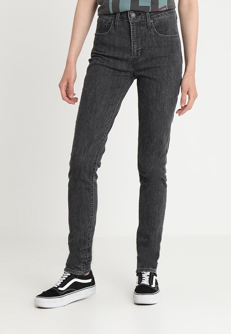 Jean gris foncé coupe slim porté avec des baskets noires et blanches par une personne debout sur un fond uni.