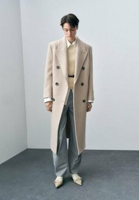 Cappotto di lana doppiopetto beige, abbinato a un maglione crema, pantaloni grigi a gamba larga e scarpe beige a punta. Bottoni neri notevoli.