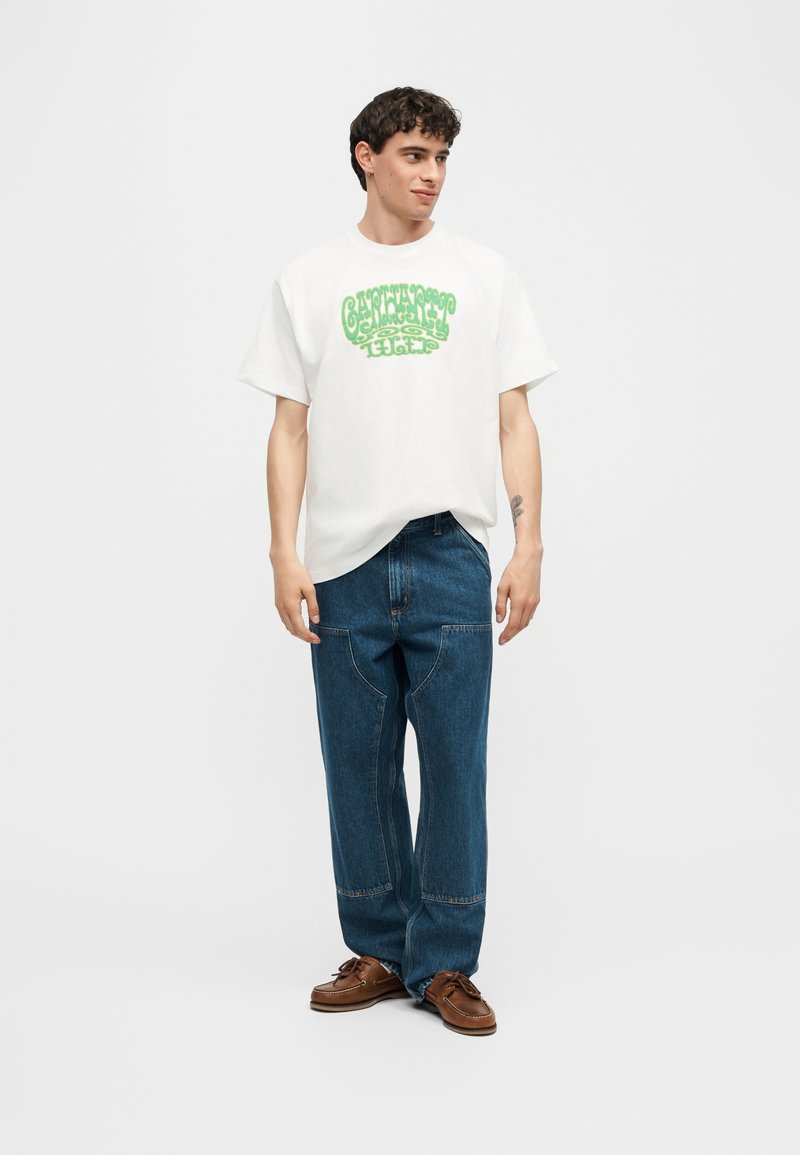 Giovane uomo con t-shirt bianca con grafica e testo verde, jeans larghi blu e scarpe di pelle marrone, in piedi su sfondo uniforme.