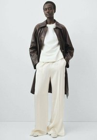 Trench coat de cuero marrón sobre una blusa blanca y pantalones de pierna ancha en crema. El conjunto presenta texturas suaves y un ajuste a medida.