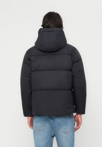 Doudoune noire avec capuche, présentant un design matelassé, des poignets élastiques et une texture lisse. Portée avec un jean en denim bleu clair.