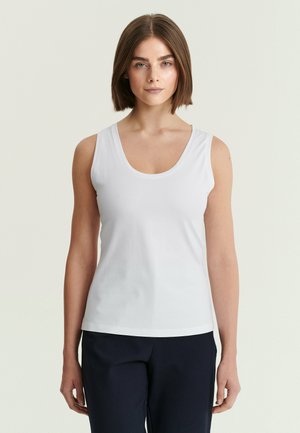 Femme aux cheveux bruns mi-longs portant un haut blanc sans manches et un pantalon foncé, debout devant un fond clair uni.