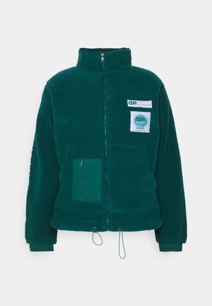 Sötétzöld, cipzáras fleece dzseki magas gallérral, elülső cipzáras zsebbel és logópatchekkel. Az alján húzózsinóros, az ujjak végén bordás mandzsettával rendelkezik.