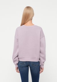 Liliowy krótki sweatshirt z obniżonymi ramionami, ribbowe mankiety i dół, zestawiony z ciemnoniebieskimi dopasowanymi dżinsami. Prosty, komfortowy design.