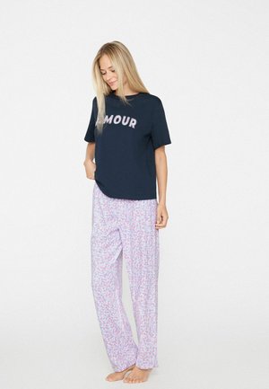 Woman wearing a navy blue t-shirt with "AMOUR" text and loose light-colored floral patterned pants, standing barefoot against a plain background.