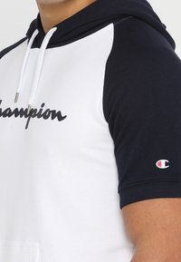 Osoba nosząca biało-czarną bluzę z krótkim rękawem z logo "Champion" na klatce piersiowej oraz małym logo "C" na rękawie.