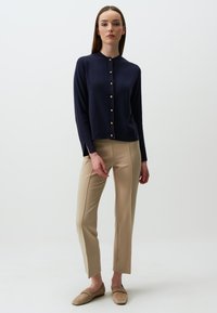 Marineblauer Strickcardigan mit goldenen Knöpfen, strukturiertem Stoff, rundem Halsausschnitt und seitlichen Schlitzen, kombiniert mit beigen, taillierten Hosen und hellbraunen Slippern.