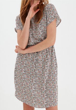 Femme aux cheveux roux portant une robe courte à manches courtes, aux genoux, à fleurs petites roses, blanches et vertes sur fond sombre.