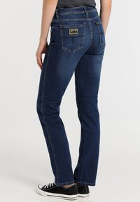 Jeans de mezclilla azul oscuro con corte recto, que cuentan con un bolsillo trasero de parche con la etiqueta "Lois", y combinados con zapatillas de lona negras.