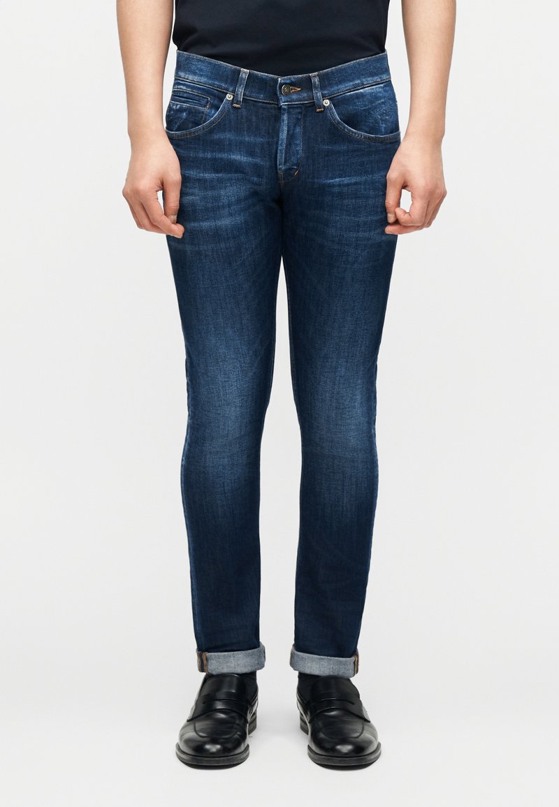 Άνδρας που φοράει σκούρα μπλε τζιν παντελόνια στενής γραμμής με γυρισμένα μπατζάκια και μαύρα δερμάτινα παπούτσια, στέκεται μπροστά σε λευκό φόντο.