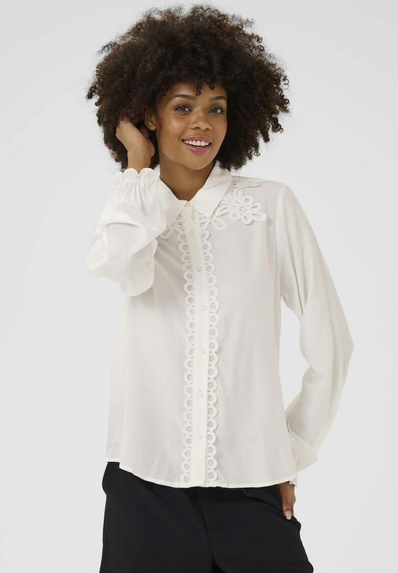 Blusa bianca a maniche lunghe con un bordo in pizzo decorativo lungo la pattina e il colletto, realizzata in un morbido tessuto trasparente e con polsini arrotondati.