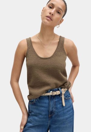 Kvinde iført brun strikket tanktop stoppet ned i højtaljede blå jeans med beige bælte, accessoriseret med gyldne hoop-øreringe og lagdelte gyldne kæder.
