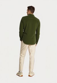 Uomo di spalle che indossa una camicia a maniche lunghe verde oliva, pantaloni beige chiaro, calze bianche e sneakers beige su uno sfondo uniforme.