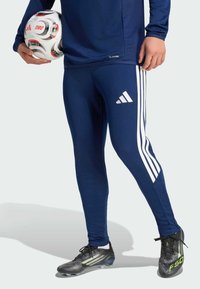 Leggings deportivos azul marino con diseño de tres rayas blancas, combinados con tacos de fútbol negros con detalles verdes y un balón de fútbol blanco.