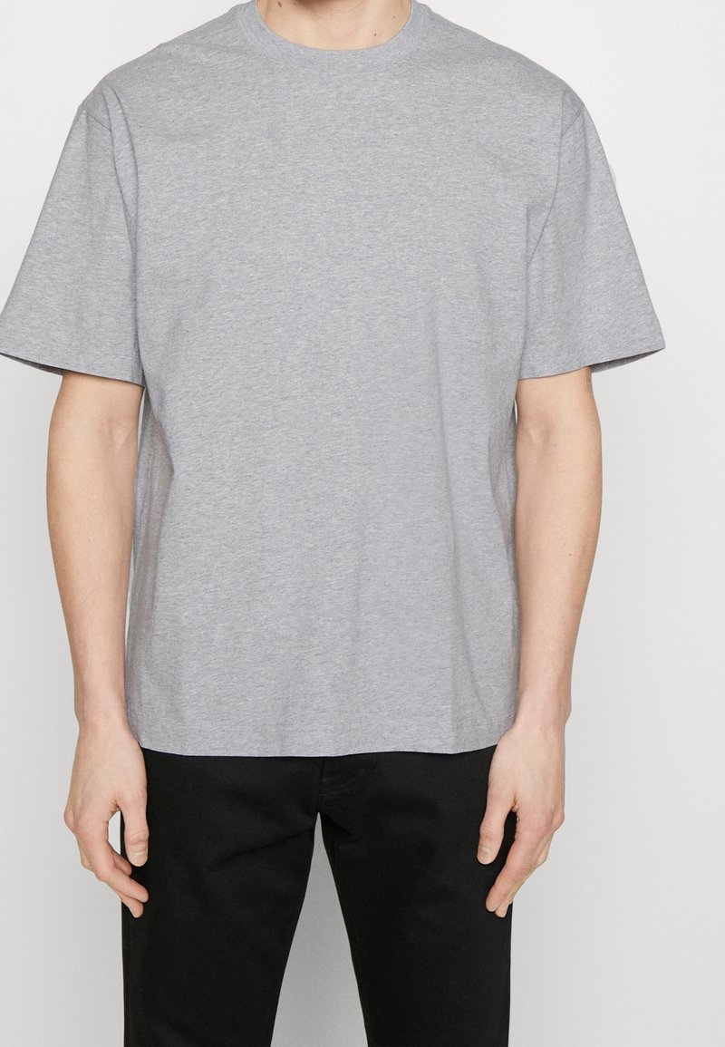 Homme portant un t-shirt à manches courtes gris clair uni et un pantalon noir, debout devant un fond blanc.