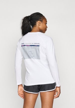 Chemise blanche à manches longues avec un graphique de panneau de départ imprimé au dos, présentant les détails des vols en noir et bleu sur un fond gris.