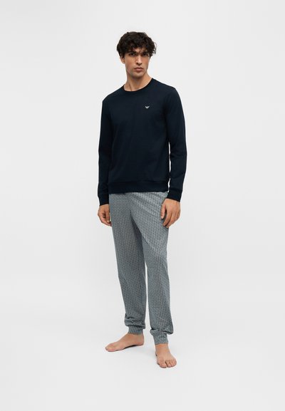 Homme debout pieds nus, portant un haut à manches longues noir et un pantalon de pyjama gris à motifs sur un fond clair uni.