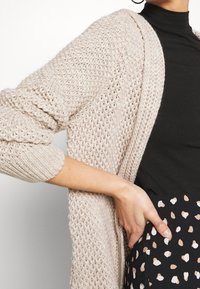 Jasnobeżowy, otwarty sweter z długimi rękawami, o teksturowanym wzorze. Nosi się go na czarną bluzkę, w zestawieniu z wzorzystą spódnicą.