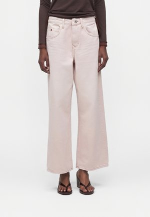 Άτομο με ανοιχτό ροζ παντελόνι ψηλής μέσης και φαρδιά μπατζάκια, σκούρο καφέ μακρυμάνικη μπλούζα και καφέ σανδάλια με τακούνι, στέκεται μπροστά σε λευκό φόντο.