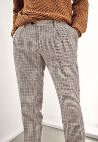 Personne portant un pull marron texturé et un pantalon à carreaux taille haute, les mains dans les poches, debout contre un mur blanc.